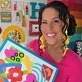 Woman in pink top holding colorful floral artwork, smiling, in a vibrant craft room